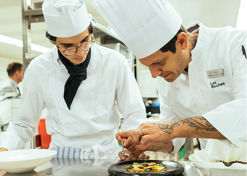 a les roches culinary student watches an instructor plate a gourmet dish